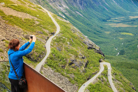 Travel concept. Tourist woman looking at Trolls Path Trollstigen or Trollstigveien winding scenic mountain road, taking picture with camera from viewing point, Norway Europeの写真素材