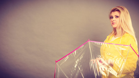 Good mood during rainy day. Happy blonde woman wearing yellow raincoat holding transparent umbrellaの写真素材