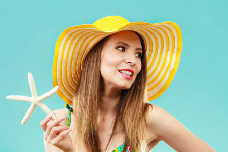 Summer holidays concept. Closeup woman in yellow hat bikini holding white shell starfish in hand on vivid blue backgroundの写真素材