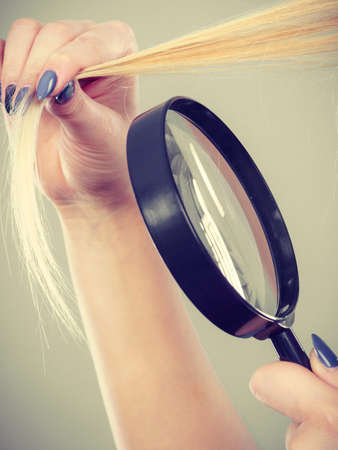 Haircare and hairstyling, bad effects of bleaching concept. Blonde woman looking at her damaged, split hair ends through magnifying glass.の写真素材