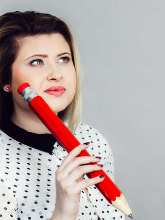 Woman confused thinking seeks a solution. Pensive thoughtful blonde student girl or young female teacher coming up with an idea, looking up, holding big red pencil. Studio shot on greyの写真素材
