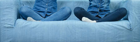 Two unrecognizable women wearing jeans matching outfit sitting on blue sofa in lotus positionの写真素材
