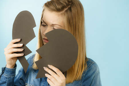 Sad young teenage woman with broken heart made of paper. Negative sad emotions, relationship problems concept. Shot on blue.の写真素材