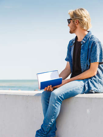Holiday, outdoor leisure time, introvert relaxation concept. Sitting hipster man wearing jeans outfit having break from reading book outside on sunny day, sea in backgroundの写真素材