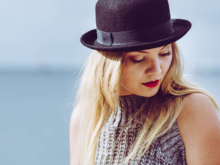 Beautiful young blonde woman wearing fedora hat, long warm sweater jumper posing next to sea beach.の写真素材