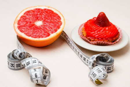 Concept of making choice: healthy low-calorie or unhealthy high-calorie food, slimming or fattening. Grapefruit and cake cupcake with measuring tapeの写真素材