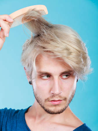 Hairdo coiffure hairstyle concept. Handsome young fashion model combing his colored blonde hair using wooden comb tool, stylish haircutの写真素材