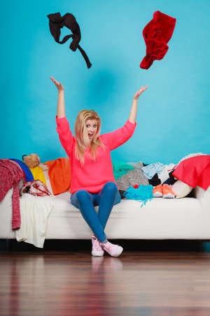 Clothing dilemmas concept. Shocked woman sitting on messy couch throwing clothes above head.の写真素材