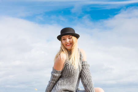 Beautiful young blonde woman wearing fedora hat and sweater jumper posing on sky background.の写真素材