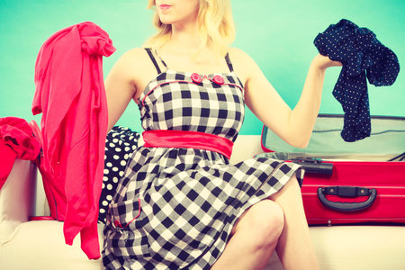 Packing problems, necessary things during the trip concept. Woman sitting on sofa, getting ready for vacation, choosing clothes to pack into suitcaseの写真素材