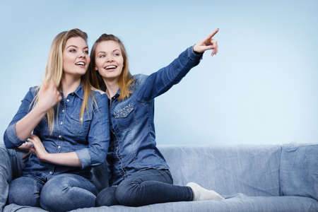 Friendship, human relations concept. Two happy women friends or sisters wearing jeans shirts sitting on sofa pointing somewhere.の写真素材