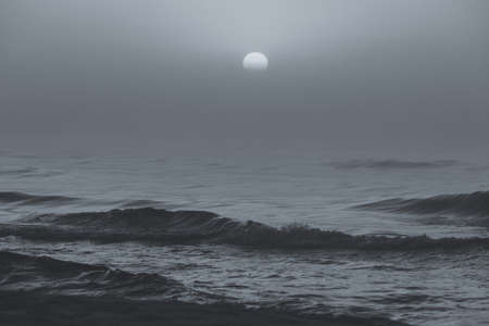 Black and white shot of sun behind fog or clouds during sunset by beach.の写真素材