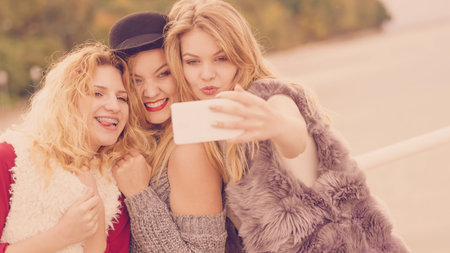 Three females taking selfies, having fun outdoor using smart phone to take photo.