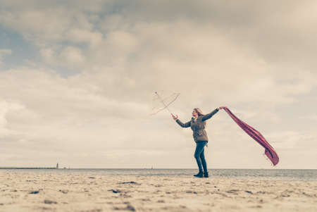 Happiness, enjoying weather, feeling great concept. Woman jumping with red scarf and transparent umbrella on beach near sea, sky with clouds in backgroundの写真素材