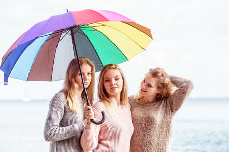 Three pretty young women friends under colorful umbrella parasol. Fashionable females wearing sweaters spending time outdoor.の写真素材