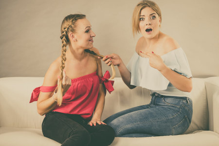 Pretty blonde shocked woman pointing at her female best friend braided hair