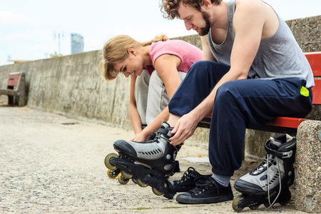 Young people friends putting on roller skates outdoor. Woman and man on bench.