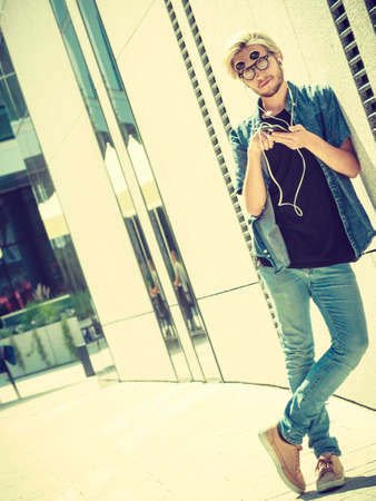 Men fashion, technology, urban style clothing concept. Hipster guy standing on city street wearing jeans outfit and weird sunglasses listening to music and looking at phoneの写真素材