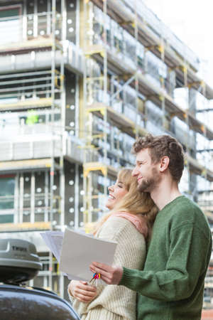 Real estate and family concept - young couple on front of new big modern house construction site with blueprint project keys dreaming about homeの写真素材