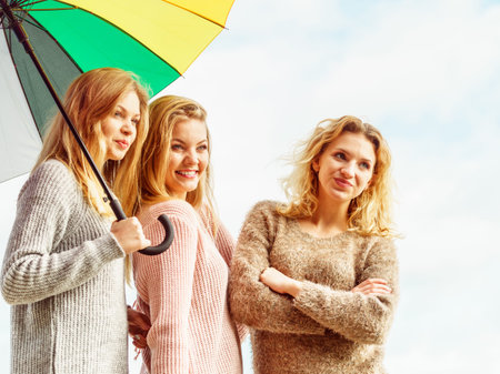 Three pretty young women friends under colorful umbrella parasol. Fashionable females wearing sweaters spending time outdoor.の写真素材