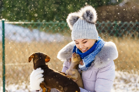 Young woman having fun during winter. Female playing with her two small purebreed dogs puppies while snow is snowingの写真素材
