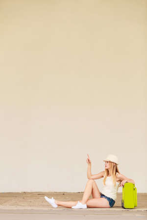 Travel, adventure, teenage journey concept. Woman wearing denim shorts, white top and sun hat suitcase holding suitcase on wheels sitting and pointing at somethingの写真素材