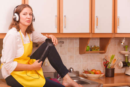 Preparing food concept. Listening music. Thoughtful worried housewife cook chef with earphones wearing yellow apron sitting at home.の写真素材