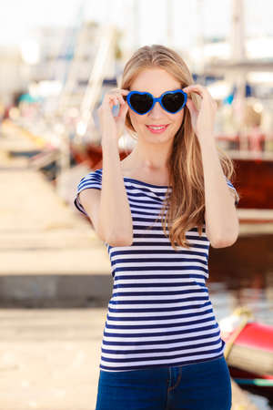Travel tourism and people concept. Fashion blonde girl with blue heart shaped sunglasses in marina against yachts in portの写真素材