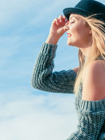 Beautiful young blonde woman wearing fedora hat and sweater jumper posing on sky background.の写真素材