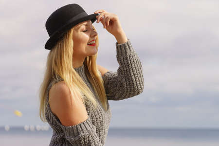 Beautiful young blonde woman wearing fedora hat and sweater jumper with holes on shoulders posing outdoorの写真素材