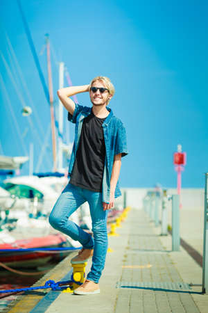 Vacation, relaxation concept. Young fashionable blonde man standing near harbor, relaxing and enjoying beautiful, sunny weather. Outdoor shotの写真素材
