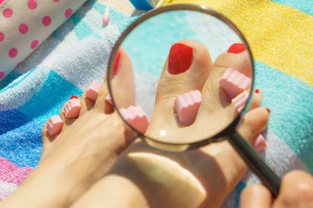 Woman looking at her fresh made pedicure through magnifying glass on towel. Female having toes nails painted with red nail polish.の写真素材