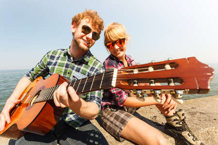 Happy young man hipster playing guitar for woman by sea ocean. Handsome guy and pretty girl in sunglasses relaxing on summer vacation.の写真素材