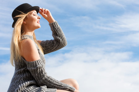 Beautiful young blonde woman wearing fedora hat and sweater jumper posing on sky background.