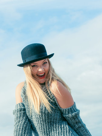 Beautiful young blonde woman wearing fedora hat and sweater jumper posing on sky background.