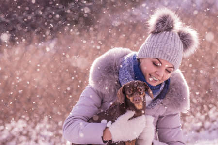 Young woman having fun during winter. Female playing with her small purebreed dachshund dog puppy while snow is snowingの写真素材