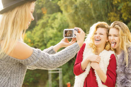 Three females friends having fun during outdoor photo session. Woman taking pictures of two during warm autumn weather.の写真素材