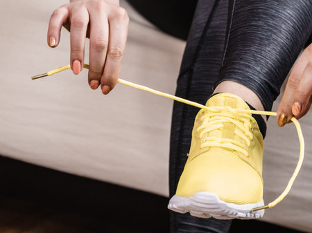 Woman in sportswear sitting on sofa indoor getting ready for exercises. Tying up shoes