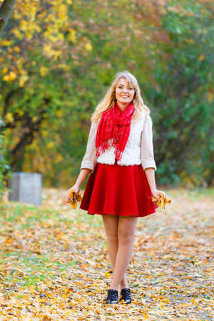 Season, happiness and people concept. Young fashionable blonde woman in full lehgth walking in autumnal park on sunny day holding leaves in handの写真素材
