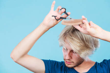 Style and fashion. Young trendy male hairstylist barber with new idea of look changing. Blonde guy with scissors and wooden comb creating hairdo coiffure, on blueの写真素材