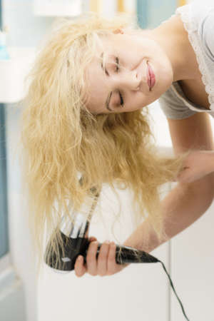 Positive woman using hair dryer on her blonde hairdo. Haircare, hairstyling concept.の写真素材