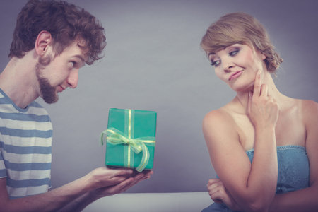 Couple sitting on couch at home. Young man giving offended woman gift box, girl refusing to accept...