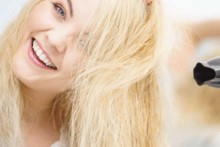 Positive woman using hair dryer on her blonde hairdo. Haircare, hairstyling concept.の写真素材