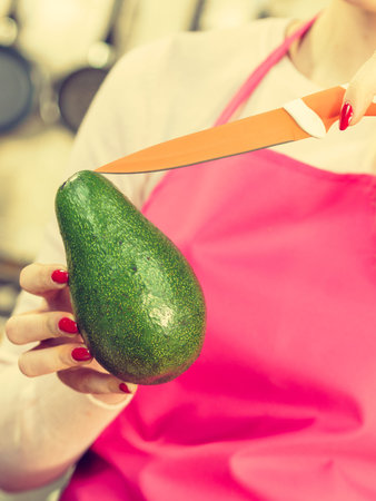 Woman cutting, preparing green vegetable, delicious avocado using kitchen knife. Female wearing...