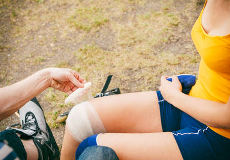 Sport injury. Couple of skaters outdoor. Young woman suffering from leg pain after taking a fall on the asphalt, man is helping bandaging her injured kneeの写真素材