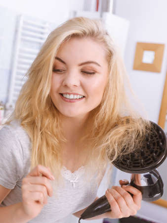 Haircare. Beauty long haired blonde woman drying hair in bathroom. Smiling girl blowing wind on wet head using hairdryer, doing curls with diffuser nozzle.の写真素材