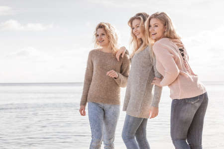 Three fashionable women wearing sweaters during warm autumnal weather spending their free time on sunny beach. Fashion models outdoor having fun.の写真素材