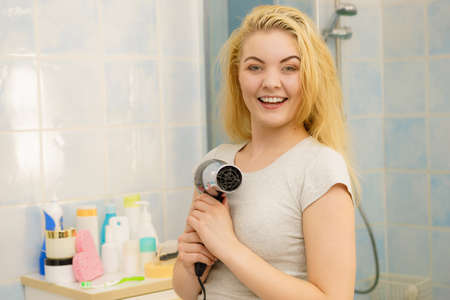 Positive woman using hair dryer on her blonde hairdo. Haircare, hairstyling concept.の写真素材