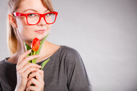 Beauty feminine casual leisure concept. Passionate lady in glasses. Young blonde girl smelling holding red tulip flower tenderly.の写真素材