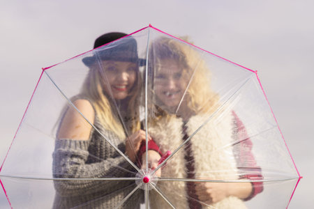 Two fashionable women wearing stylish outfits holding transparent umbrella spending their free time...